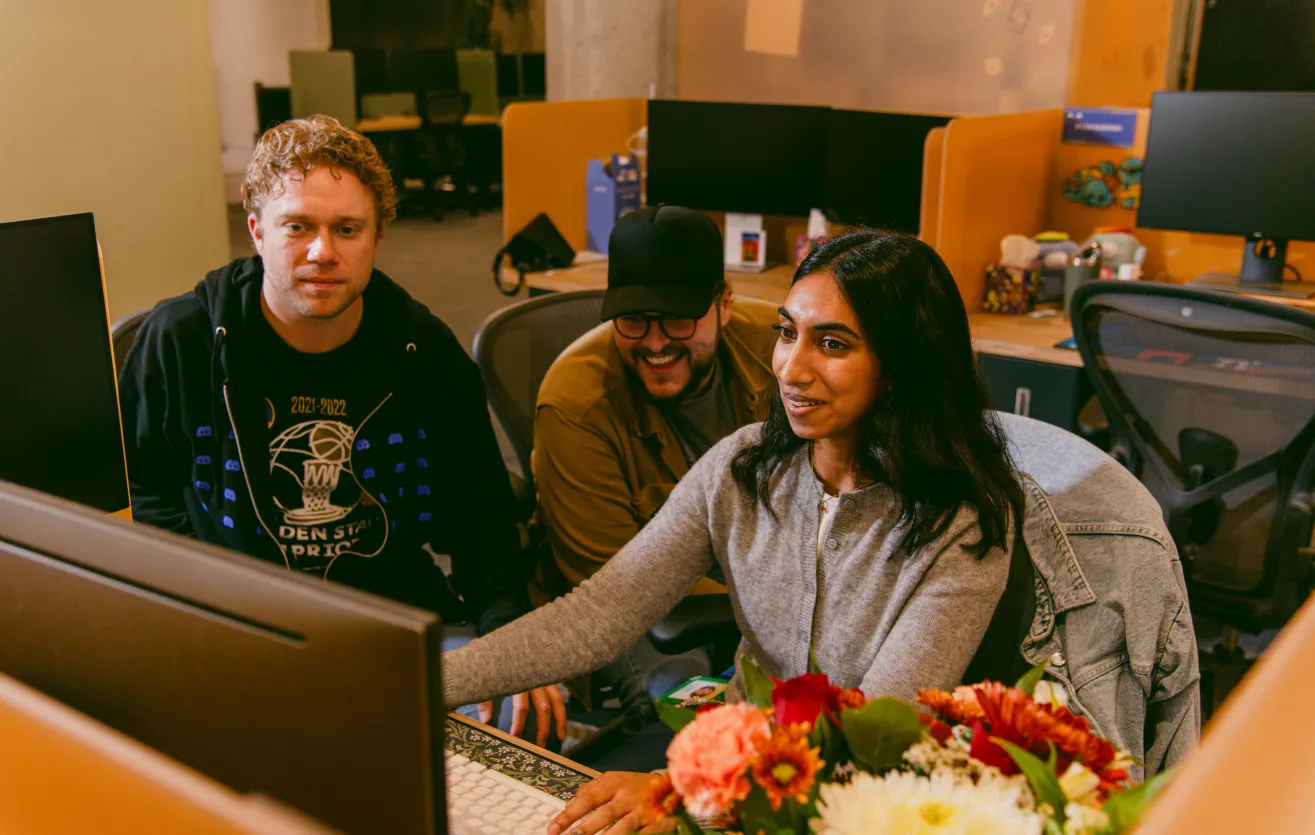 Three Discord employees are collaborating at one workstation, deep in what appears to be a productive conversation. One employee is controlling the keyboard and mouse, appearing to direct the conversation. The three of them are seated near the center of the office