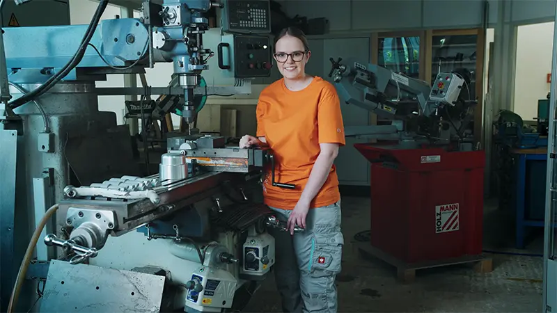 Ein weiblicher Lehrling der Integrativen Betriebe arbeitet an einer Standbohrmaschine und lächelt in die Kamera