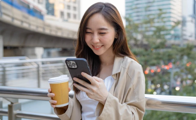 A woman smiles while looking at her phone and standing outdoors.