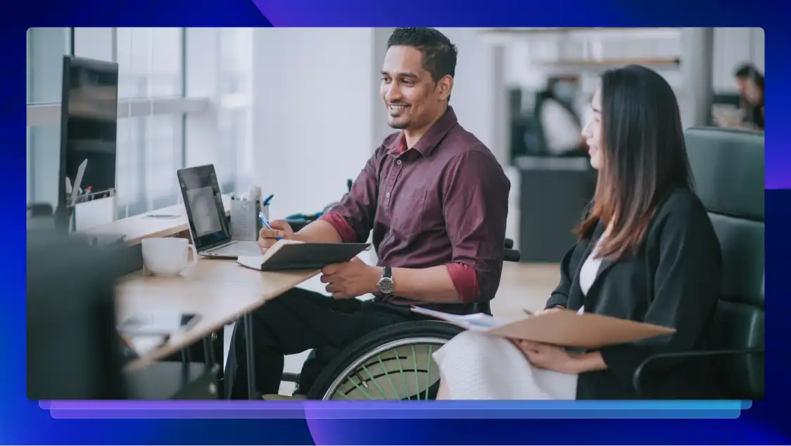 Man and woman at an office desk smiling and taking notes in front of a computer. The man is in a wheelchair.