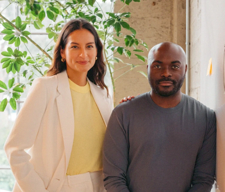 Woman in white suit and man in grey sweater facing the camera in warm and brightly lit office